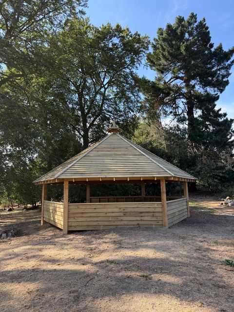 The outdoor classroom from across the field, its hexagonal shingle roof standing among mature trees.