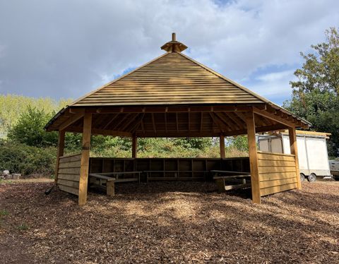 An open-sided hexagonal outdoor classroom with a steep timber roof, half-height boarded walls and a bark chipping floor.