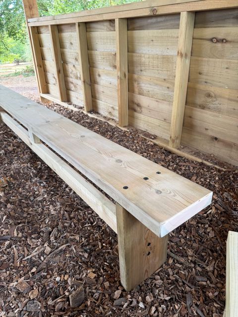 Close view of a bench, showing the planed timber seat, its supporting legs and the boarded wall behind.