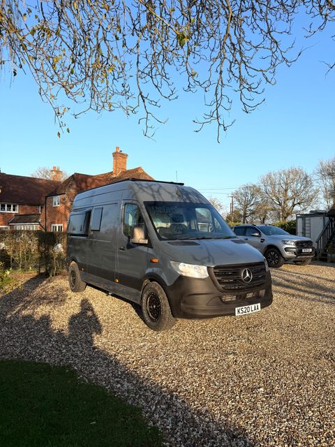 A dark grey Mercedes Sprinter campervan with a high roof and black alloy wheels, parked on gravel in front of a tiled cottage.