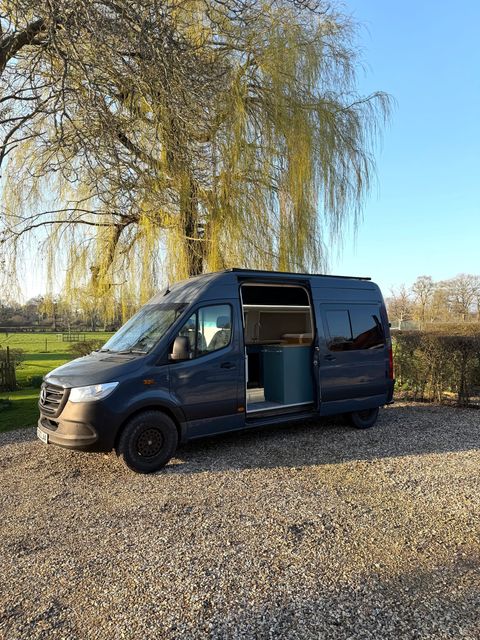 The campervan with its side door slid open beneath a willow tree, showing the fitted interior.