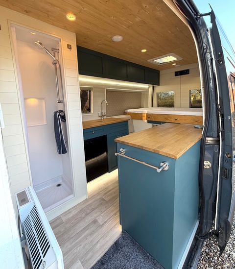 Looking into the campervan through the open side door: shower cubicle on the left, teal kitchen units with oak worktops, bed across the back.