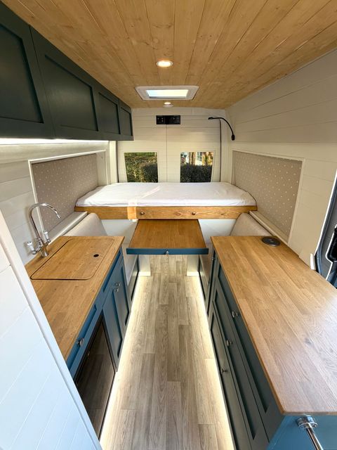 The interior looking towards the back: oak worktops down both sides over teal cabinets, a drop-down table in the middle, and a bed across the rear under a rooflight.