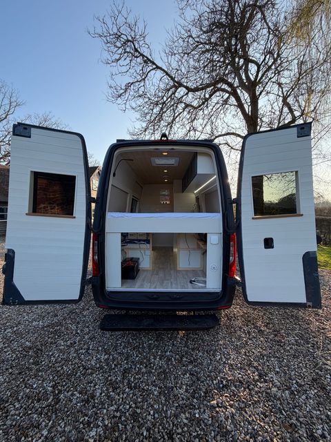 The rear doors open on the campervan, panelled in white inside with windows set into each door, showing the bed and the interior beyond.