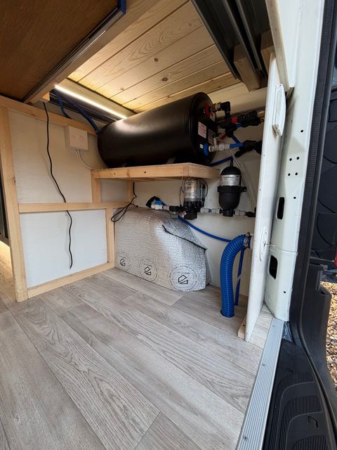 The plumbing bay under the bed, with a black water tank on a timber shelf above the pump, filter and insulated pipework.